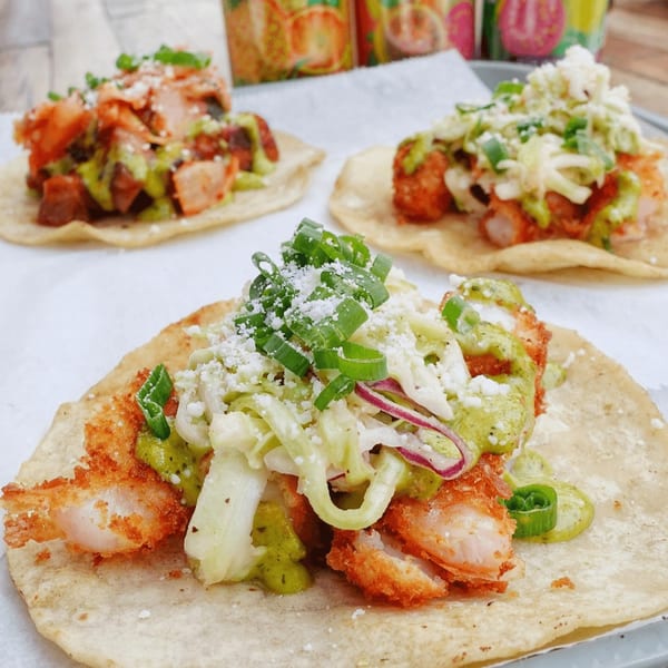 Three vibrant tacos with various toppings, including fried shrimp, green sauce, and shredded cabbage.