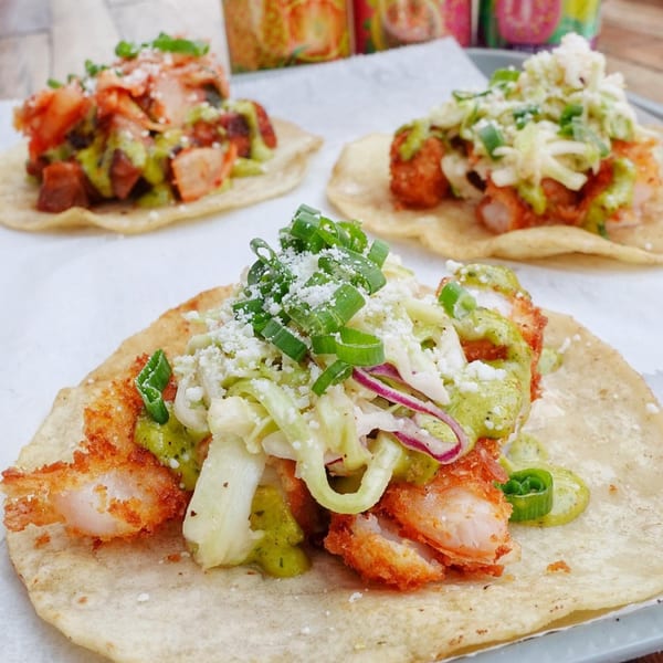 Three vibrant tacos with various fillings, including fried shrimp, cabbage, and green sauce, on a white surface.