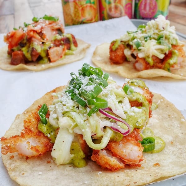 Three vibrant tacos with various toppings, including fried shrimp, green sauce, and shredded cabbage.