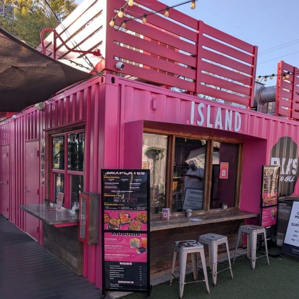 A vibrant pink shipping container restaurant, "Island Faka's Island Grill," with outdoor seating and menus.