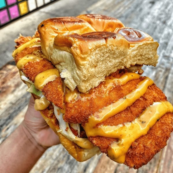 Hand holding a large fried chicken sandwich with cheese sauce, lettuce, and Hawaiian rolls.