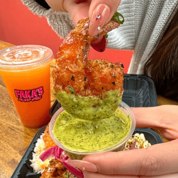 A hand dips a crispy fried shrimp into a small cup of vibrant green sauce, with a drink in the background.
