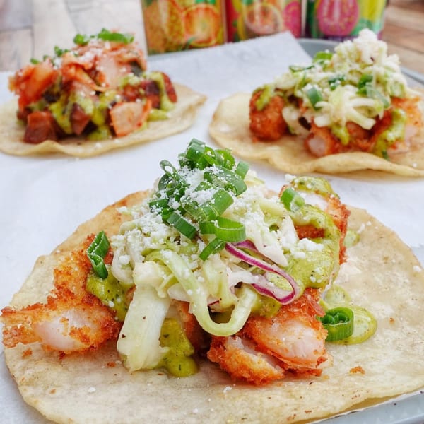 Three vibrant tacos with fried shrimp, green sauce, cabbage, and cheese on a white surface.