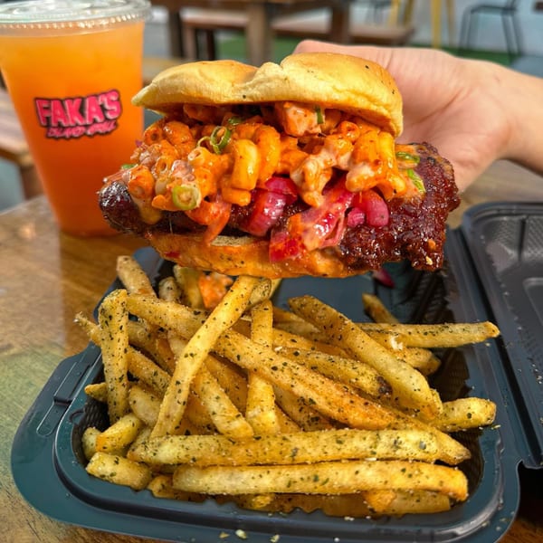 A hand holds a Faka's burger with mac and cheese, red onions, and sauce, next to seasoned fries and a drink.
