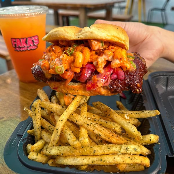 Hand holding a Faka's burger with mac and cheese, pickled onions, and a side of fries.
