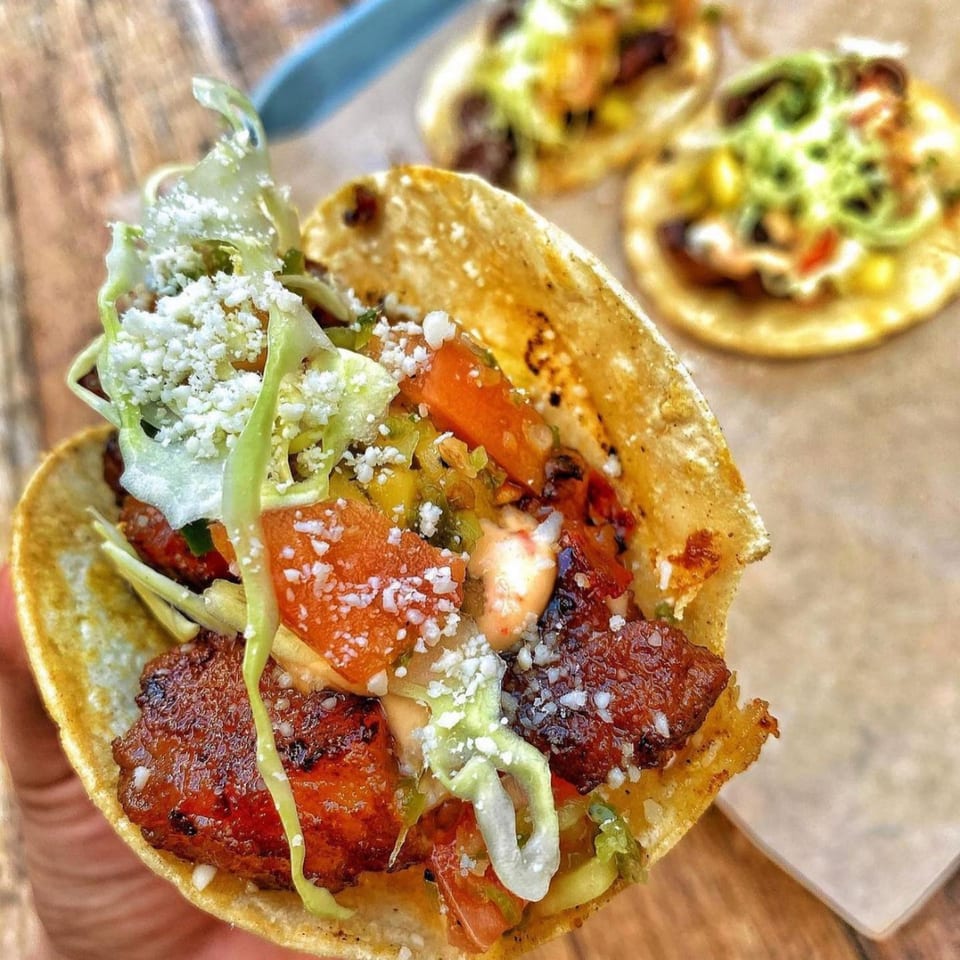 A hand holds a taco filled with meat, lettuce, tomatoes, and cheese, with two more tacos in the background.