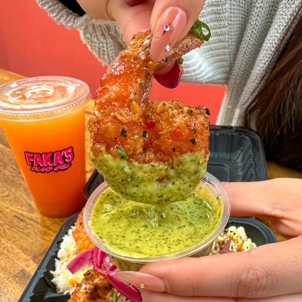 A hand dips a crispy fried shrimp into a small container of green sauce, with a drink in the background.