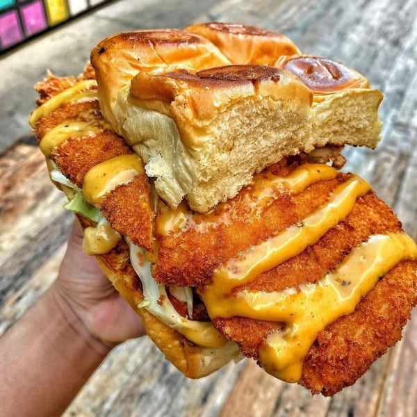 Hand holding a large fried chicken sandwich with cheese sauce and shredded lettuce on a bun.